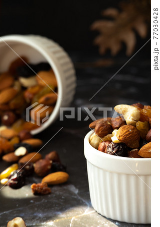 Mix of delicious assorted nuts and dried fruit in white cups on a table, sunny morning. Healthy gourmet vegetarian natural snack. Mix of delicious assorted nuts and dried fruit in white cups on a table, sunny morning. Healthy gourmet vegetarian natural snack. 77030428