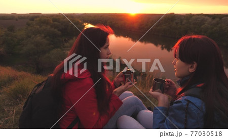 Mugs in hand at sunset on a hike. Girls hold mugs of tea in their hands at dawn. Drink hot drinks while traveling on the background of the sky in the glare. Adventures of life. Girlfriends have a rest 77030518