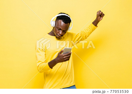 Young African man in casual attire wearing wireless headphones listening to music with mobile phone Young African man in casual attire wearing wireless headphones listening to music with mobile phone 77053530