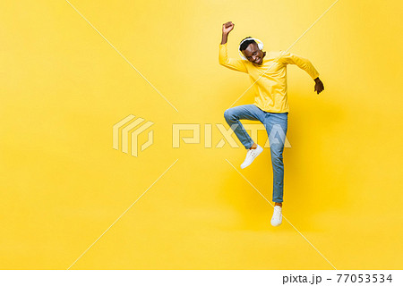 Happy energetic young African man wearing headphones listening to music and jumping with hand up Happy energetic young African man wearing headphones listening to music and jumping with hand up 77053534