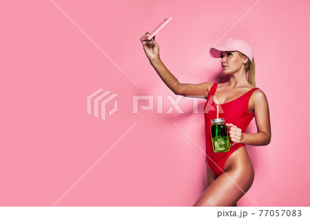 Smiling young blonde woman in red one-piece swimsuit cap doing selfie shot isolated on pink background. Smiling young blonde woman in red one-piece swimsuit cap doing selfie shot isolated on pink background. 77057083