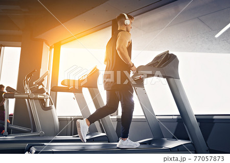 Concentrated fit man listening to music in headphones while running on treadmill in modern gym with panoramic windows. 77057873