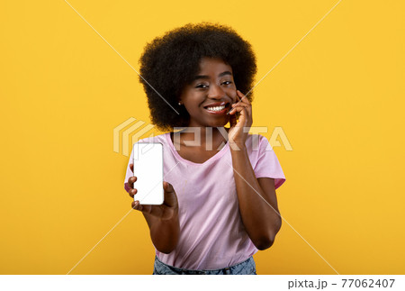 Mobile app advertising. Pretty black bushy woman showing smartphone with blank screen on yellow studio background Mobile app advertising. Pretty black bushy woman showing smartphone with blank screen on yellow studio background 77062407