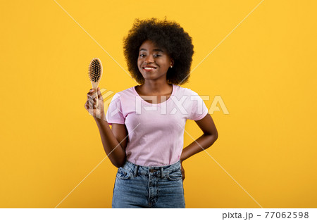 Happy african american woman holding wooden hair brush, smiling to camera over yellow studio background Happy african american woman holding wooden hair brush, smiling to camera over yellow studio background 77062598