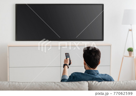 Man watching television, turning on plasma flatscreen TV-set, pointing remote control at empty TV screen on white wall 77062599
