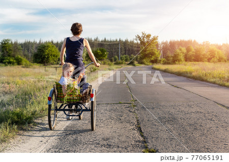 Young adult caucasian mom enjoy having leisure fun riding bicycle with cute adorable blond daughter holding wild field flower at scenic rural country road on bright sunny day. Countryside vacation Young adult caucasian mom enjoy having leisure fun riding bicycle with cute adorable blond daughter holding wild field flower at scenic rural country road on bright sunny day. Countryside vacation 77065191