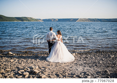 groom and bride stand in embrace on rocky bank near river in summer 77067028