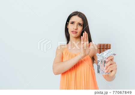 Beautiful young woman with stop gesture refuses eating chocolate bar Beautiful young woman with stop gesture refuses eating chocolate bar 77067849