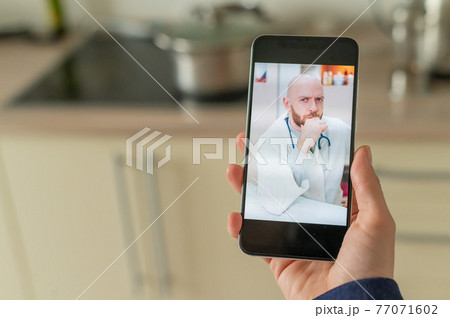 A serious bearded doctor communicates with a patient via a video call on a smartphone. Online medicine concept. Woman in the kitchen takes therapist remote consultation on mobile. A serious bearded doctor communicates with a patient via a video call on a smartphone. Online medicine concept. Woman in the kitchen takes therapist remote consultation on mobile. 77071602
