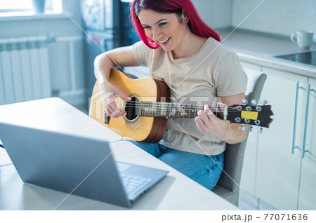 A woman sits in the kitchen during a remote acoustic guitar lesson. A girl learns to play the guitar and watches educational videos on a laptop A woman sits in the kitchen during a remote acoustic guitar lesson. A girl learns to play the guitar and watches educational videos on a laptop 77071636