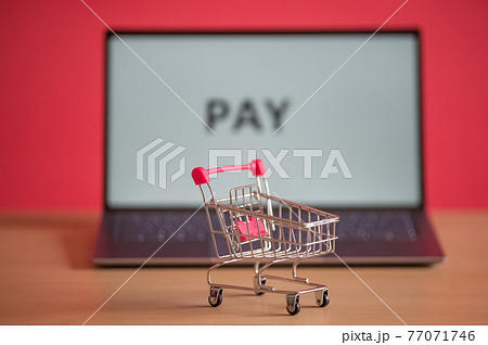 Mini shopping trolley stands in front of a laptop on a pink background. Online shopping concept. The word pay on a computer screen. Remote purchase of products and things on the Internet. 77071746