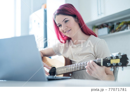 A woman sits in the kitchen during a remote acoustic guitar lesson. A girl learns to play the guitar and watches educational videos on a laptop 77071781