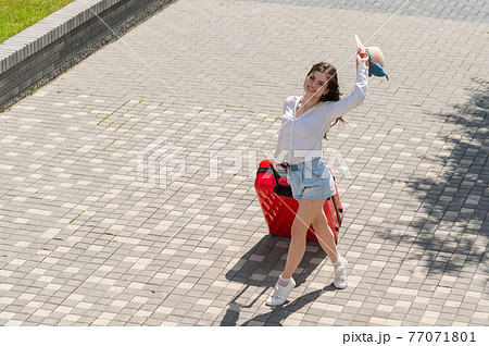 Happy caucasian young woman in hat and shorts holding a large red suitcase at an open age Happy caucasian young woman in hat and shorts holding a large red suitcase at an open age 77071801