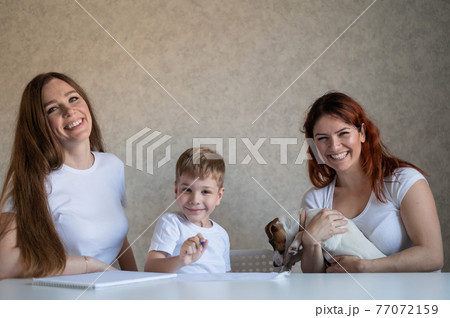 Happy family stays at home. Two women help the boy do school homework. Lesbian couple sitting at the table with their son and a cheerful puppy. Same-sex marriage with a child. Distance learning. Happy family stays at home. Two women help the boy do school homework. Lesbian couple sitting at the table with their son and a cheerful puppy. Same-sex marriage with a child. Distance learning. 77072159