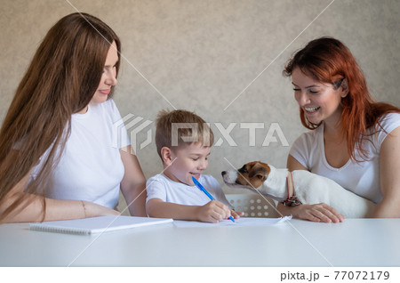 Happy family stays at home. Two women help the boy do school homework. Lesbian couple sitting at the table with their son and a cheerful puppy. Same-sex marriage with a child. Distance learning. 77072179