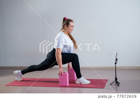 Young fat caucasian woman lunges on a sports mat. Charming plus size girl in sportswear is doing fitness exercises with an online trainer on the phone against a white background Young fat caucasian woman lunges on a sports mat. Charming plus size girl in sportswear is doing fitness exercises with an online trainer on the phone against a white background 77072639