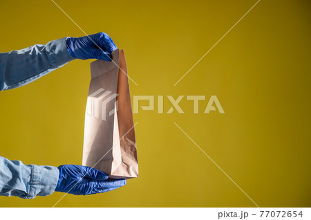 Closeup of female hands in gloves and a denim shirt. Delivery man passes an empty paper bag on a yellow background. Craft packaging for takeaway snack. Antimicrobial protection. 77072654