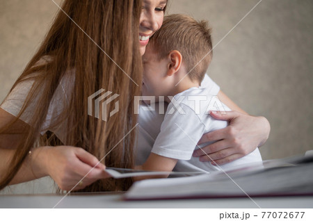 A middle-aged Caucasian woman hugs a little son and shows a photo album. A little boy sits on his mothers lap and looks at old photographs. Pleasant memories. Close family relationships. A middle-aged Caucasian woman hugs a little son and shows a photo album. A little boy sits on his mothers lap and looks at old photographs. Pleasant memories. Close family relationships. 77072677