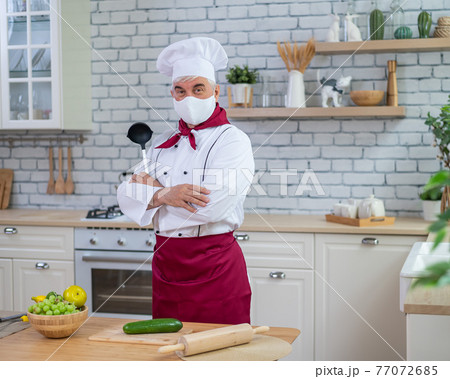 An elderly male chef wearing a mask in the kitchen crossed his arms over chest An elderly male chef wearing a mask in the kitchen crossed his arms over chest 77072685