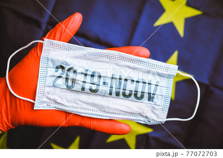The inscription on the protective mask - coronavirus 2019-ncov is held by a doctor in red gloves against the background of the EU flag. The concept of the spread of the disease epidemic. 77072703