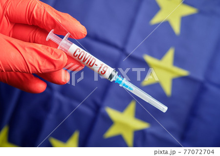 Doctor holds a syringe with the inscription virus COVID19 on the background of the EU flag. The concept of a virus epidemic in countries. Doctor holds a syringe with the inscription virus COVID19 on the background of the EU flag. The concept of a virus epidemic in countries. 77072704