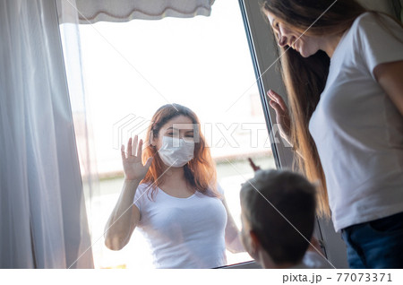 A masked woman is standing outside by a window plane, waving to her sister and nephew who are staying at home. Social distance among families in quarantine. Coronavirus. The girl at the window pane. A masked woman is standing outside by a window plane, waving to her sister and nephew who are staying at home. Social distance among families in quarantine. Coronavirus. The girl at the window pane. 77073371