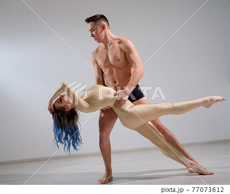 Acrobatic couple perform number on a white background. A duet of gymnasts rehearsing a performance with support. A man and a very flexible woman are dancing. 77073612