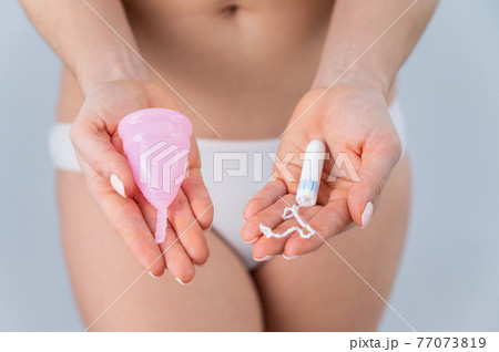 A faceless woman in cotton panties holds a pink menstrual cup and tampon on a white background. Various hygiene products during menstruation 77073819