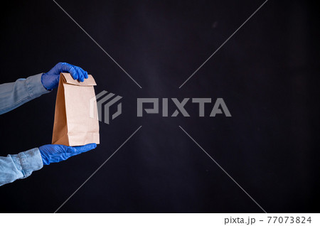 Female hands in a blue glove holds a brown paper bag on a black background. Safe food delivery to your home. A courier in a denim shirt holds out a craft cardboard bag to a customer. Female hands in a blue glove holds a brown paper bag on a black background. Safe food delivery to your home. A courier in a denim shirt holds out a craft cardboard bag to a customer. 77073824