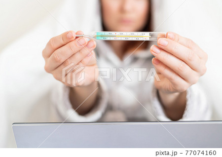 A woman in pajamas with a fever shows a mercury thermometer at the camera. Faceless blurry girl with fever is sitting on the bed. 77074160