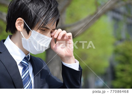 目元を触っているマスクをしてスーツを着た男性 77086468