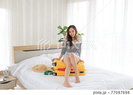 young Asian woman in summer casual clothes is smilling and relaxing mood sitting on a yellow suitcase in her bedroom. It is a time to go to travel in a summer time. Happy tourist woman with yellow young Asian woman in summer casual clothes is smilling and relaxing mood sitting on a yellow suitcase in her bedroom. It is a time to go to travel in a summer time. Happy tourist woman with yellow 77087698