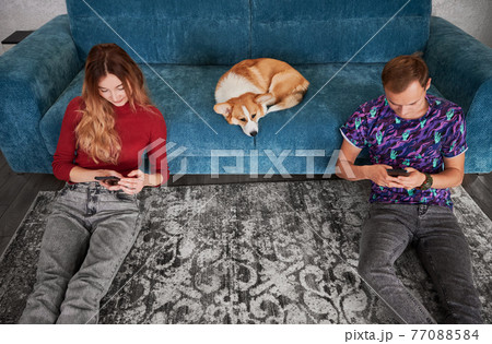 Young couple with smartphones and cute Corgi on couch. 77088584