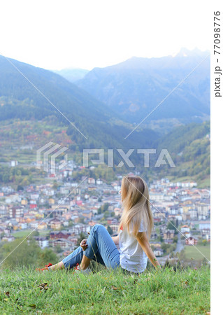Young woman sitting in mountains, little village in background. 77098776