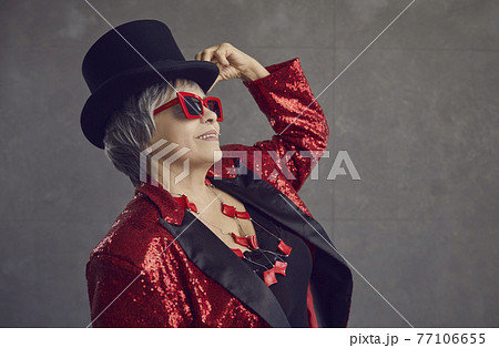 Happy youthful looking senior woman in top hat, cool glasses and sequin jacket posing in studio 77106655