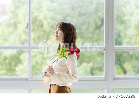 窓辺で薔薇を2輪持ち立っている若い女性 窓辺で薔薇を2輪持ち立っている若い女性 77107239