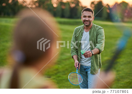 Happy father holding badminton racket and shuttlecock while playing with his little daughter outdoors in the park on a summer day 77107801