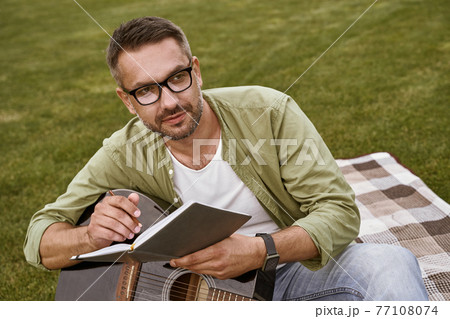 Male musician working in park. Young focused man wearing eyeglasses holding acoustic guitar and composing a song while sitting on a green grass outdoors Male musician working in park. Young focused man wearing eyeglasses holding acoustic guitar and composing a song while sitting on a green grass outdoors 77108074