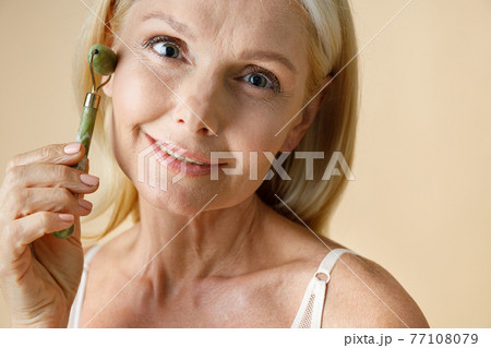 Close up portrait of mature blonde woman with glowing skin smiling at camera while using jade facial roller for skin care and beauty treatment, posing isolated over beige background Close up portrait of mature blonde woman with glowing skin smiling at camera while using jade facial roller for skin care and beauty treatment, posing isolated over beige background 77108079