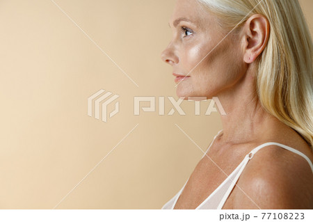 Side view portrait of pretty mature blonde woman in white underwear looking away, standing isolated over beige background 77108223