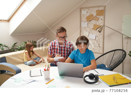 Dad helping his son with school homework, boy learning online while his little sister sitting on sofa on the background Dad helping his son with school homework, boy learning online while his little sister sitting on sofa on the background 77108256
