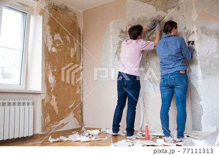 Young couple removing old wallpaper from walls preparing for flat renovation. 77111313
