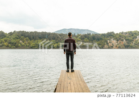Man standing lonely on a wooden pier by the lake with a view of a nature Man standing lonely on a wooden pier by the lake with a view of a nature 77113624