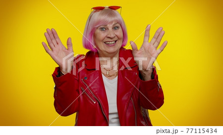 Happy friendly elderly woman waves hand palm in hello gesture welcomes someone positive emotions 77115434