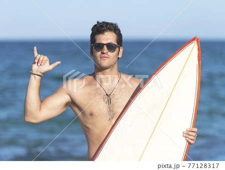 Surfer guy happy with surf surfing smiling doing hawaiian shaka hand sign for fun during surf session in ocean waves on beach vacation. 77128317