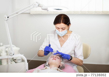 Female cosmetologist wearing face mask and medical gloves, applying cream or foam on a girl's face to clean her skin. A cosmetology procedure in a beauty salon for skin acne treatment. 77136002