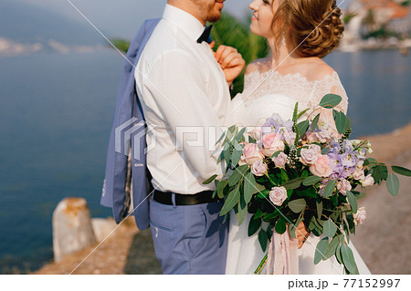 The bride and groom stand embracing on the pier, the bride holds a bouquet in her hand, close-up  77152997