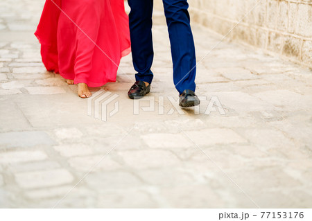 Barefoot bride in a long bright pink dress and the groom walking along a cobbled road, close-up 77153176