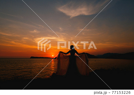 The bride at sunset stands on the seashore and holds the ends of the dress in her stretched out hands. 77153226