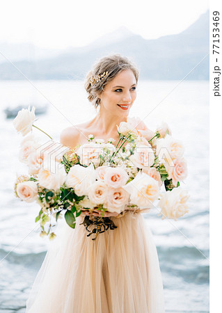 Smiling bride with a bouquet of flowers stands on the shore of Lake Como against the backdrop of mountains Smiling bride with a bouquet of flowers stands on the shore of Lake Como against the backdrop of mountains 77153469